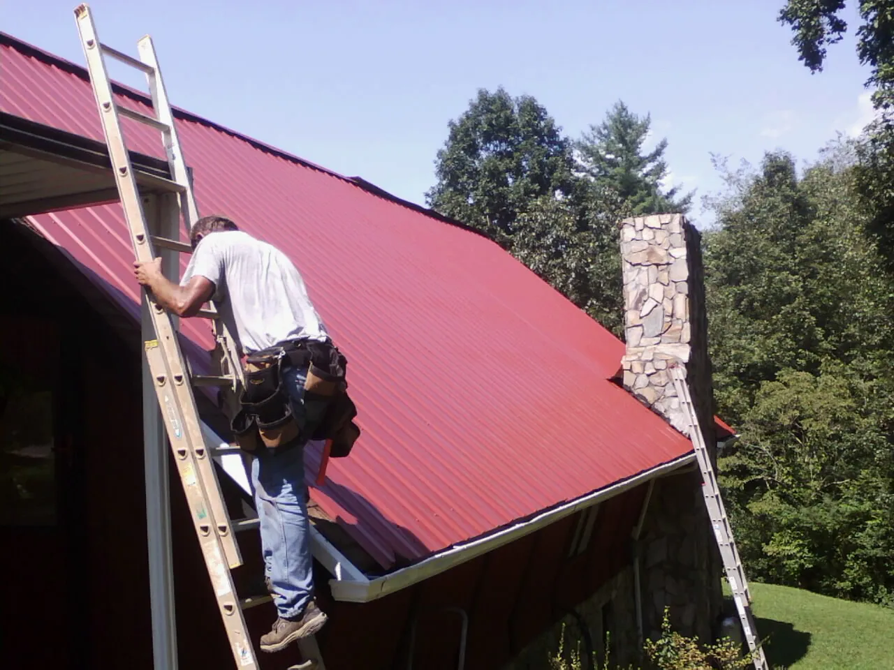 Roofer on a ladder beside a red metal roof and stone chimney — Soffit & Fascia Repair by Roofing Brothers in Hilham